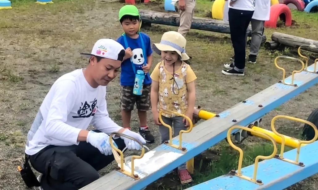 公園のシーソーを白い塗料で塗り直すボランティアと、その様子を見守る子どもたち